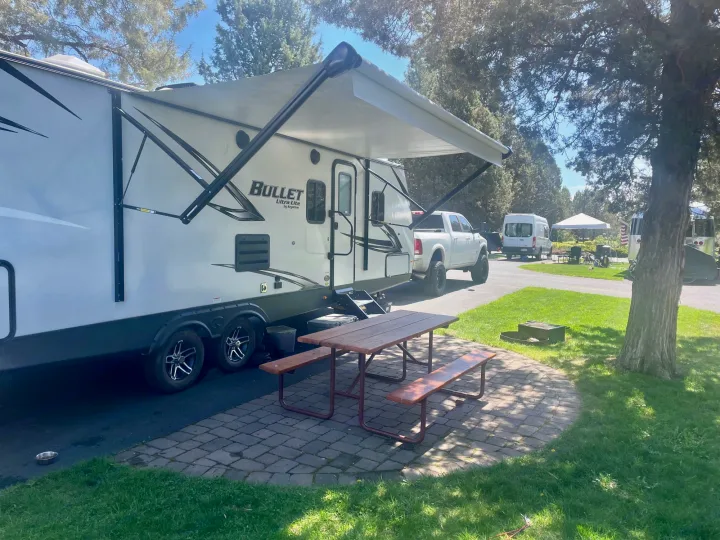 A white travel trailer RV rental, featuring an extended awning and picnic table, parked at a sunny campsite.. Keystone Bullet 2021