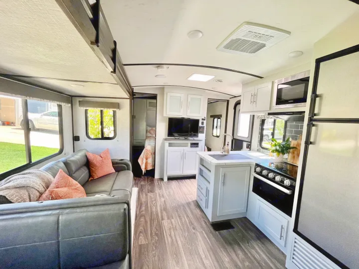 Inside a clean RV rental, featuring a gray sofa, full kitchen with oven/microwave, and wood-look floors leading to a bedroom.. Keystone Bullet 2021
