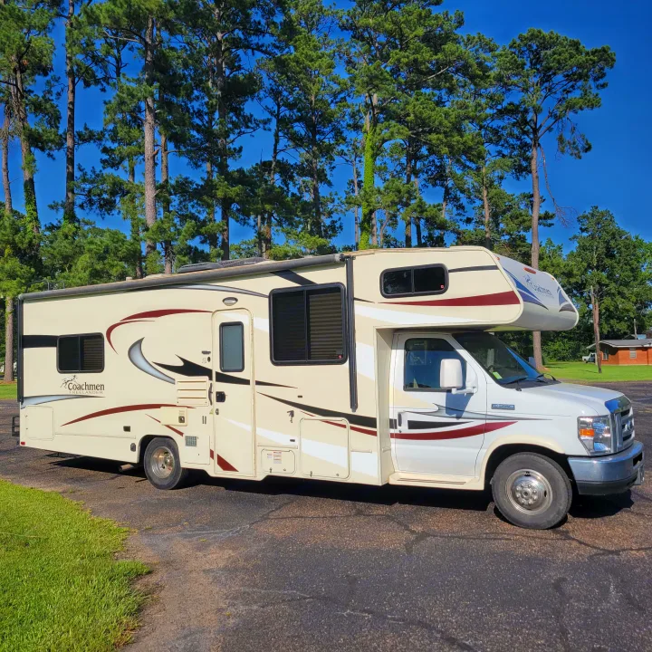 Cream & white Coachmen Class C RV rental. Parked on asphalt with tall pine trees in background.. Forest River Coachmen Freelander 2016