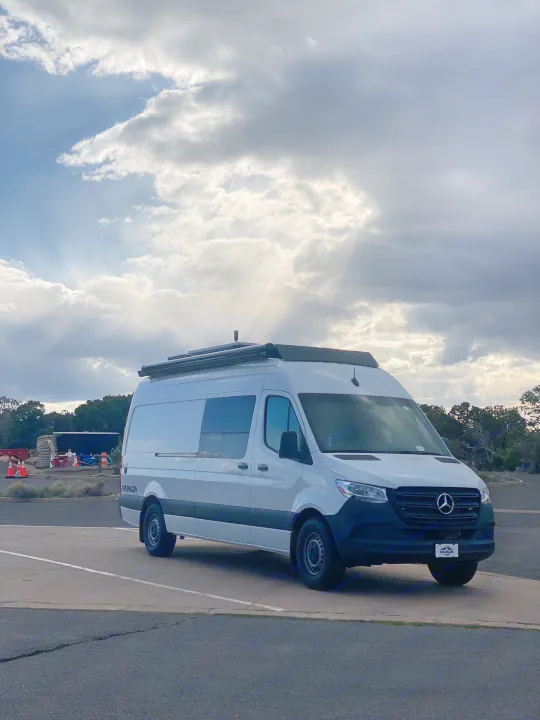 A white Mercedes-Benz Sprinter RV rental parked under a dramatic sky with sun rays. Ideal for scenic travel and adventure.. Mercedes-Benz Sprinter 2022