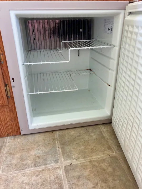 Open and empty RV mini-fridge for an rv rental, showing its interior shelves and cooling elements, with some signs of use.. Jayco Qwest 2003