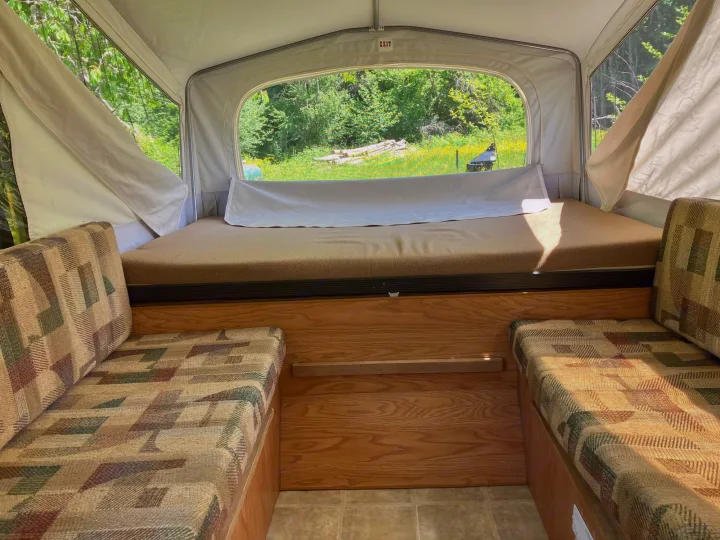 Cozy pop-up RV rental interior with patterned benches, a bed, and a scenic view of nature. Perfect for your next adventure.. Jayco Qwest 2003