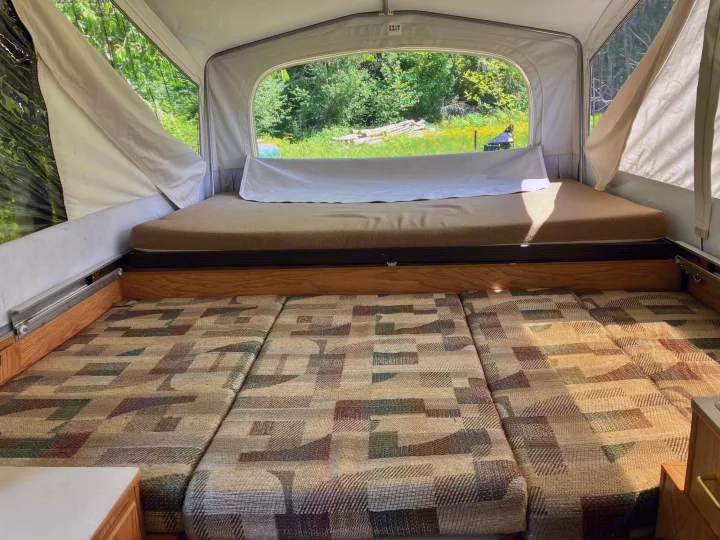 Inside a cozy pop-up RV rental: Two sleeping areas, patterned cushions, and a large window view of nature.. Jayco Qwest 2003