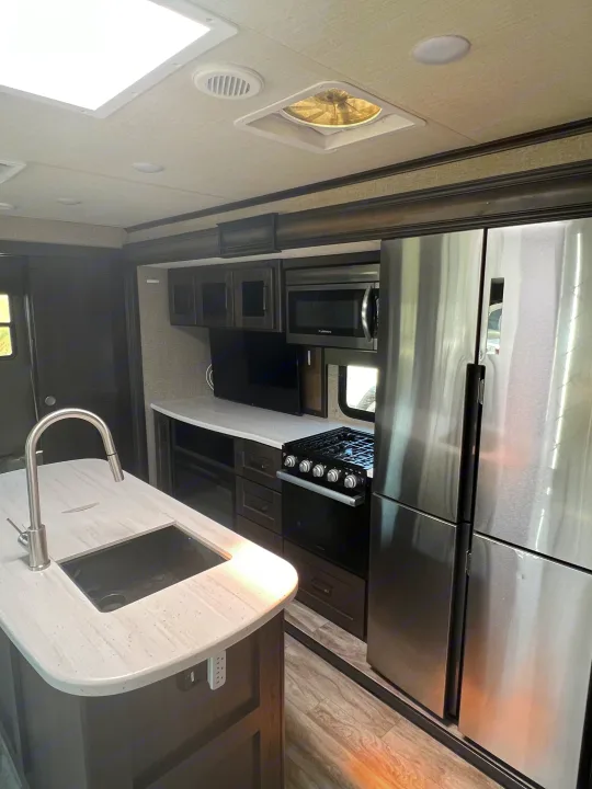Inside a modern RV rental: sleek kitchen with stainless steel appliances, island sink, full oven, fridge, & living space.. Grand Design Reflection 2022
