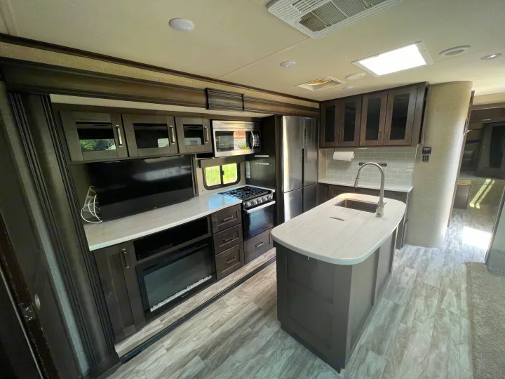 Interior view of a modern RV rental featuring a full kitchen with an island, stainless steel appliances, a television, and a fireplace.. Grand Design Reflection 2022