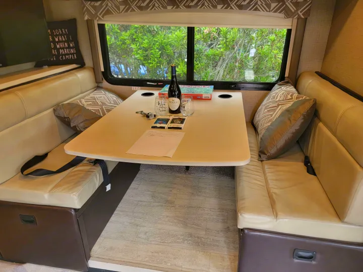 Cozy RV rental dinette setup with wine, chocolate, and Monopoly, perfect for travel relaxation.. Thor Axis 2021