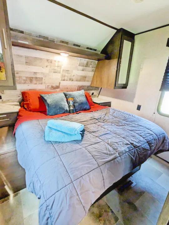 Cozy RV rental bedroom with queen bed, wood paneling, overhead cabinets, and fresh towels for a comfortable stay.. Coleman Lantern 2021