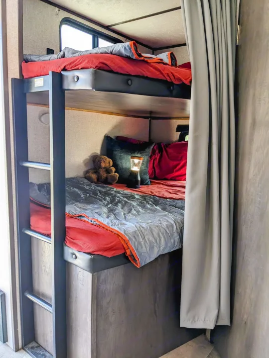 Cozy RV rental bunk beds with red sheets, gray comforters. A teddy bear and lantern sit on the lower bunk.. Coleman Lantern 2021