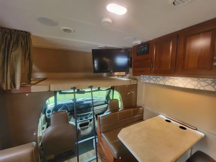 Inside a comfy **RV rental**: view of the front cab, an over-cab bunk bed, dinette with table, and overhead TV/controls.. Forest River Sunseeker 2017