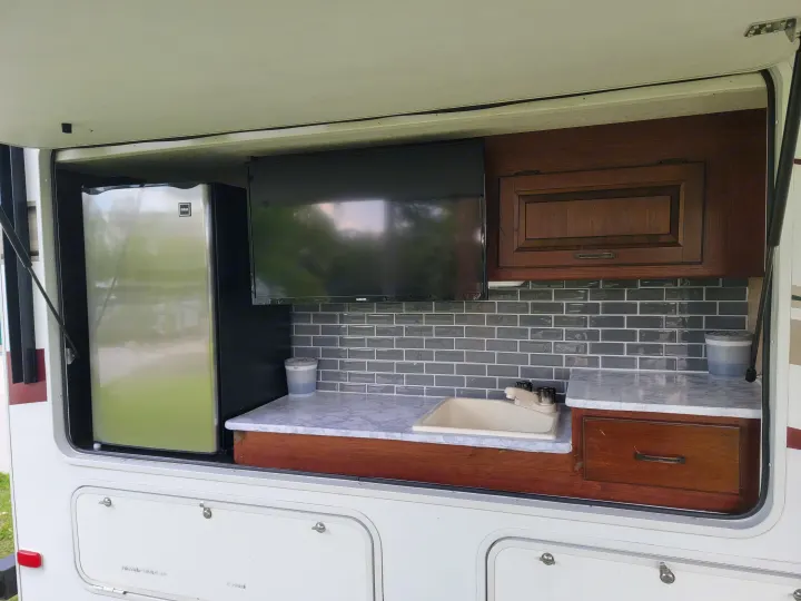 An open hatch reveals a well-equipped RV rental outdoor kitchen, complete with a fridge, TV, sink, and storage.. Forest River Sunseeker 2017