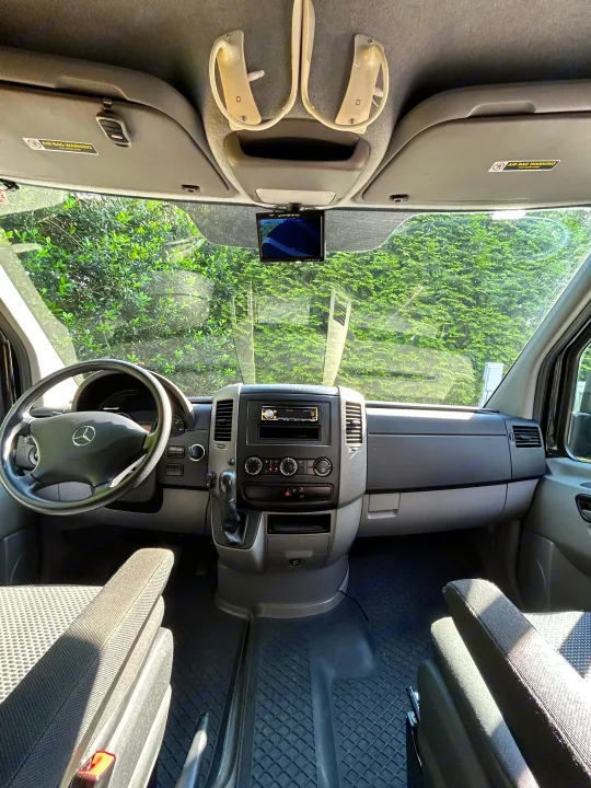 RV rental's front interior: Mercedes dashboard, steering wheel, and central console, with a clear view of green foliage outside.. Mercedes Sprinter 2012