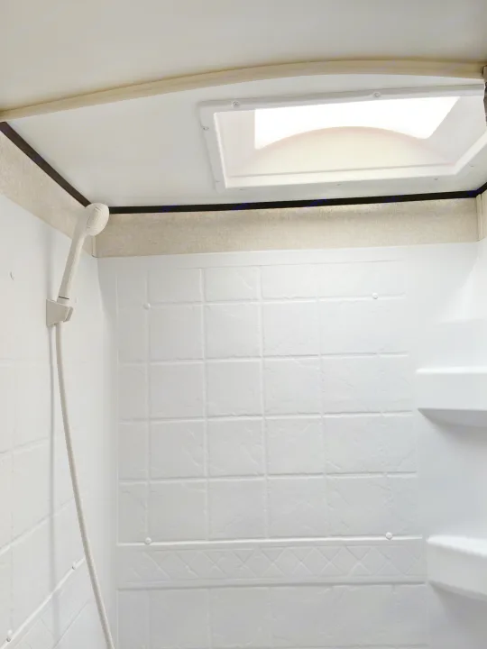 Bright RV rental shower stall: white textured walls, handheld shower head, built-in shelf, and ceiling skylight.. Jayco Jay Flight SLX 2018
