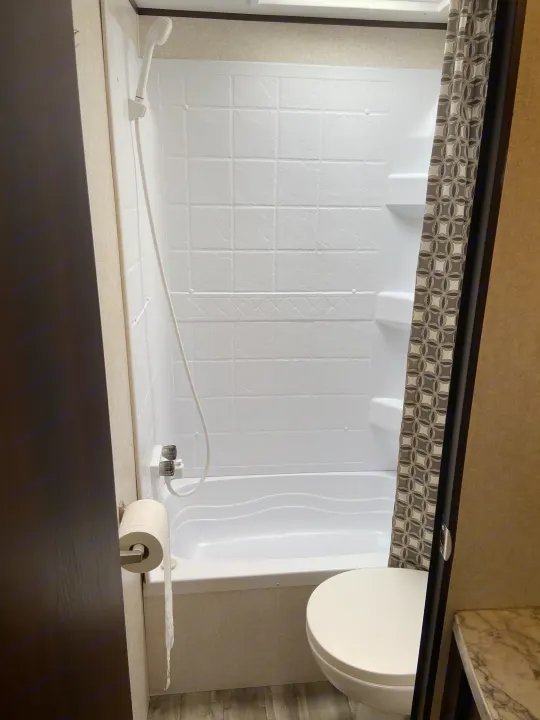 RV rental bathroom features a white shower/tub unit with a patterned curtain, toilet, and wall-mounted toilet paper holder.. Jayco Jay Flight SLX 2018