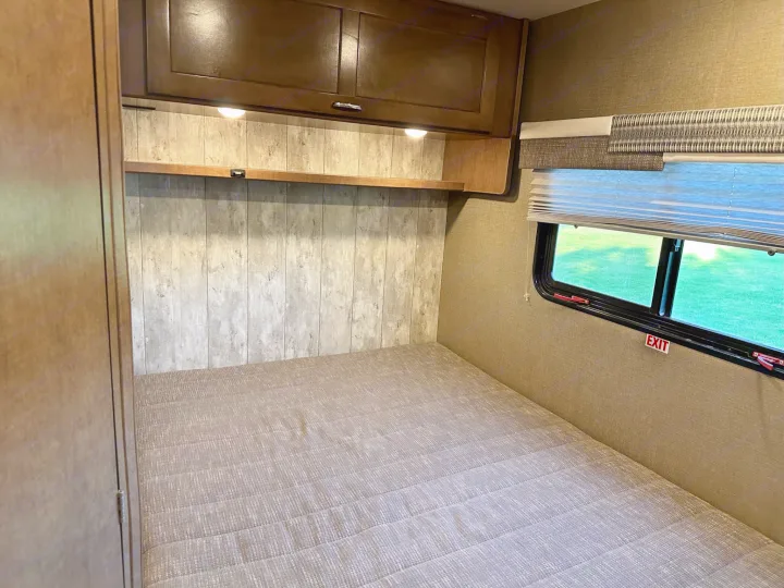 RV rental bedroom interior featuring a bed, overhead cabinets, and window with an exit sign.. Winnebago Outlook 2019