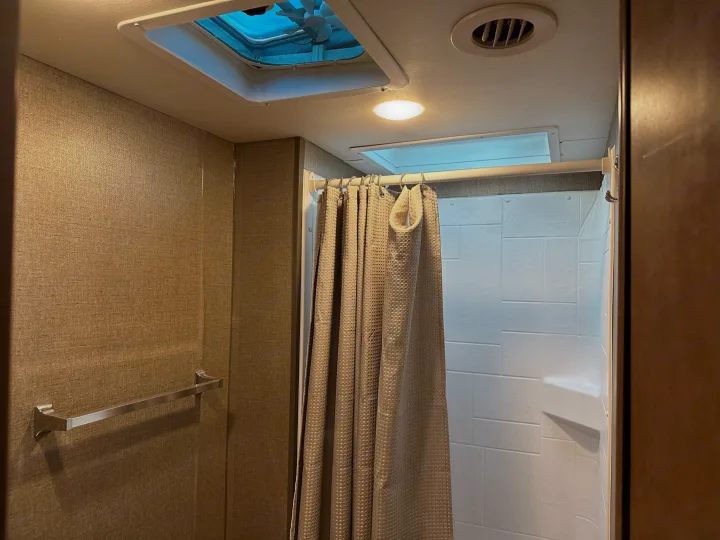A compact RV rental bathroom featuring a shower with a textured curtain, white walls, and practical overhead ventilation.. Winnebago Outlook 2019