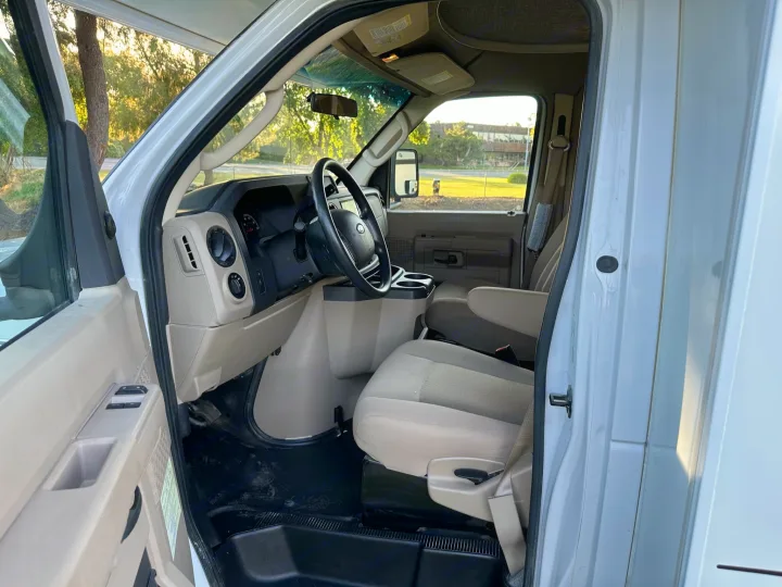 Inside an RV rental: driver's area with tan seats, dashboard, and black steering wheel; open door reveals sunny outdoor scene.. Winnebago Outlook 2019
