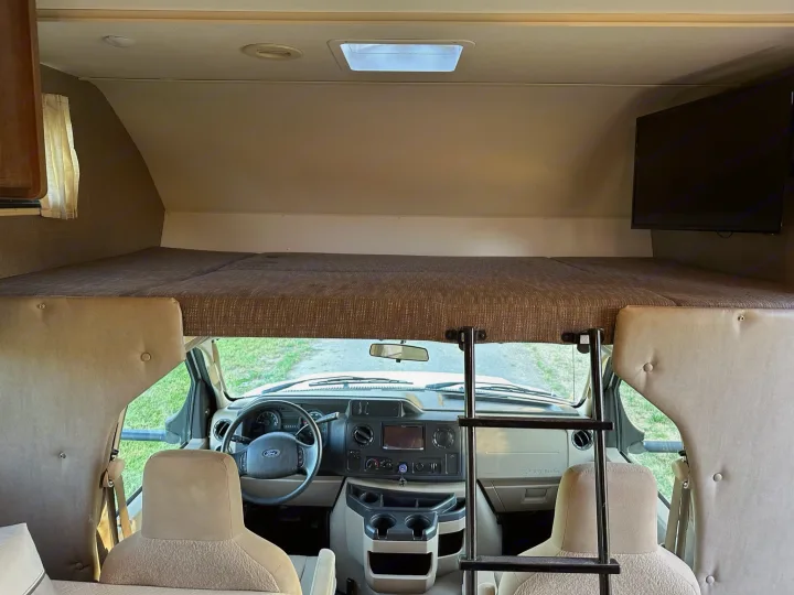 Inside an RV rental: the front cabin with Ford dashboard, and the cozy over-cab bunk bed with a TV and access ladder.. Winnebago Outlook 2019