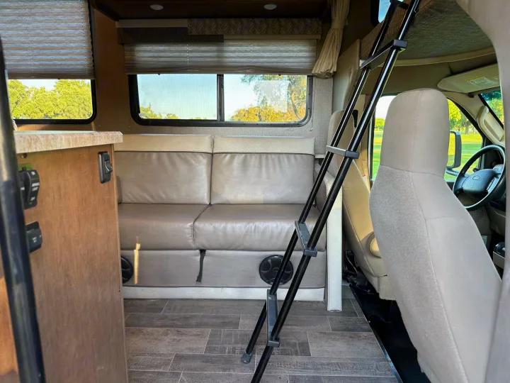 Inside an RV rental: a cozy living area with a light brown sofa, large windows, and a bunk ladder near the cab.. Winnebago Outlook 2019