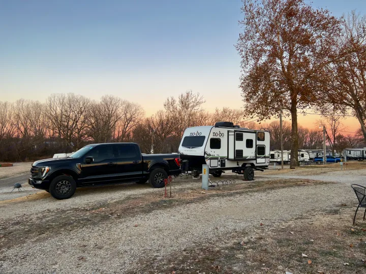 A black truck and white RV are parked at a gravel campground site. This RV rental is ready for camping.. No Boundaries 16 2023