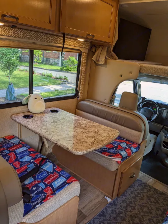 Cozy RV rental interior with a dinette booth, table, and an overhead bunk featuring a TV. Ready for travel adventures.. Thor Freedom Elite 2017