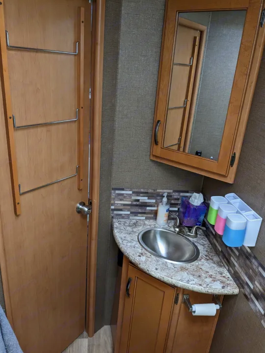 Compact RV rental bathroom featuring a sink vanity, mirror, and door-mounted towel/clothes hooks for storage.. Thor Freedom Elite 2017