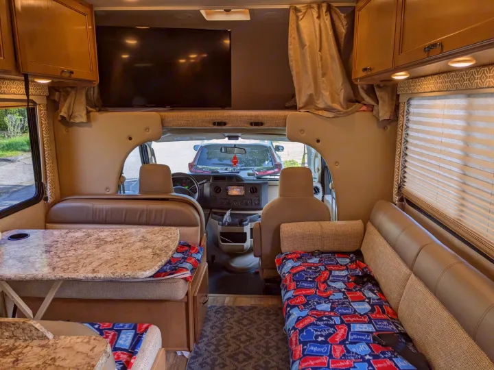 Spacious RV rental interior featuring a dinette, sofa, and overhead bunk with TV, leading to the front cockpit area.. Thor Freedom Elite 2017