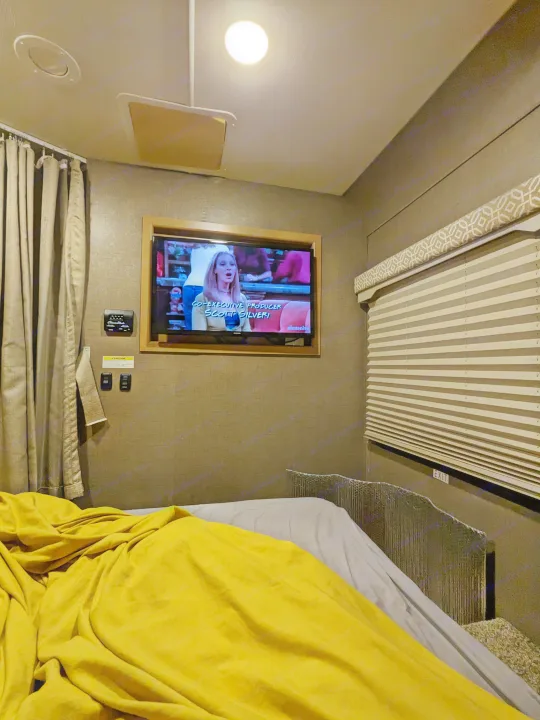 Cozy RV rental bedroom with a yellow-covered bed, TV showing 'Friends', window blinds, and controls on the wall.. Thor Freedom Elite 2017