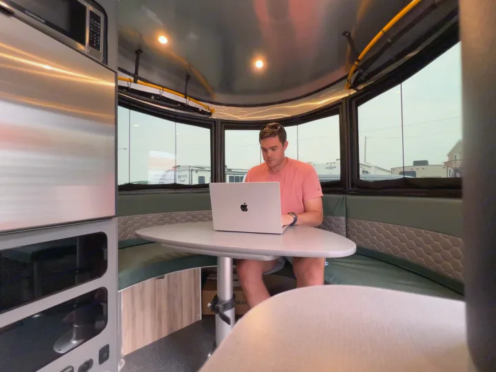 Man works remotely from a modern RV rental's interior, featuring a sleek desk, seating, and large windows.. Airstream Basecamp 2023