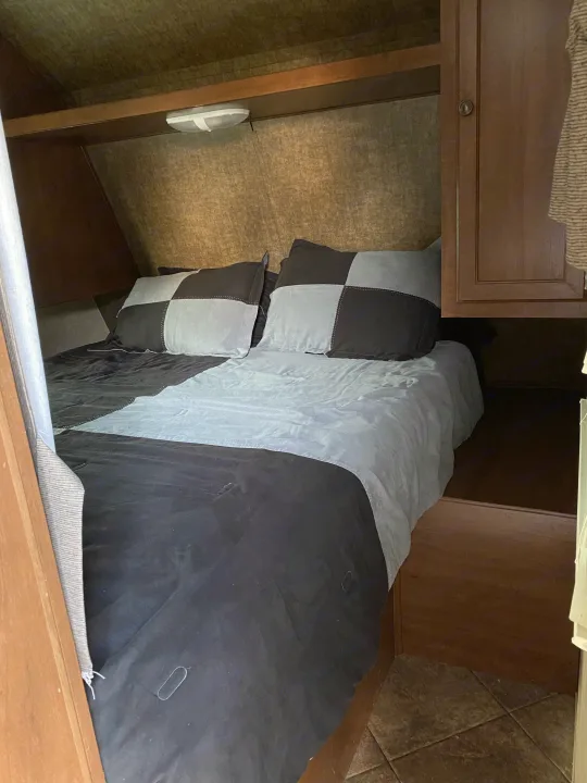 Cozy RV rental bedroom with a checkered bed, wooden cabinet, and shelf, ideal for comfortable travel.. Heartland RVs 30LX 2014