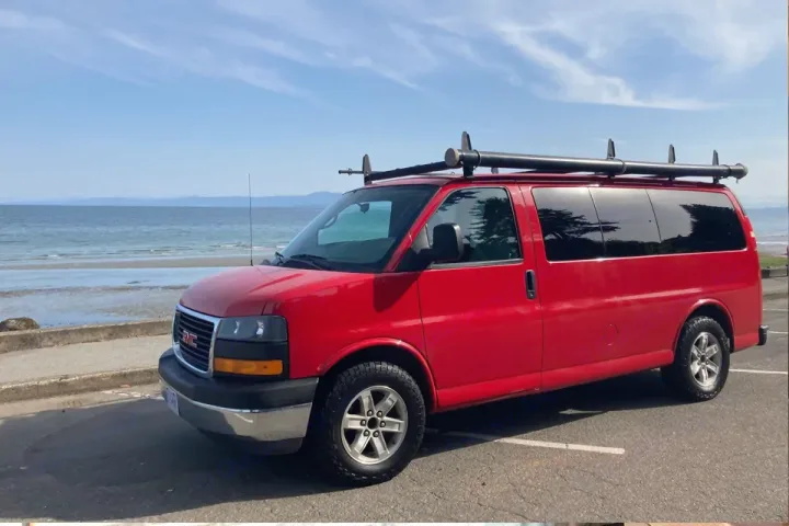 Red GMC van, available as an RV rental, parked at a scenic beach. Explore coastal adventures.. Gmc Savanna 2010