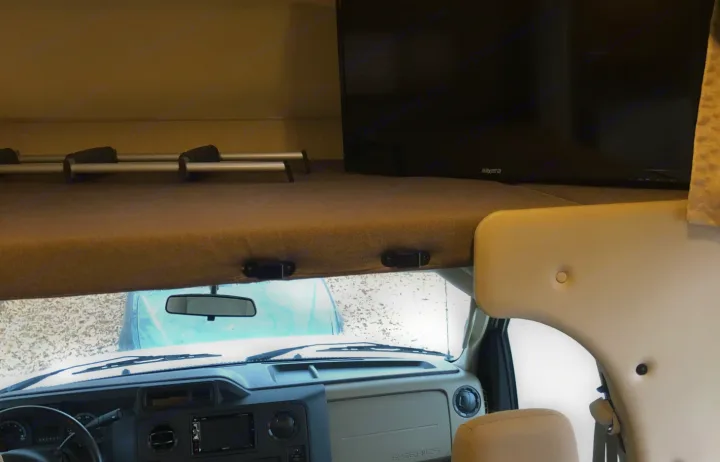 This RV rental interior shows the cab-over bunk with a TV above the dashboard and windshield.. Thor Four Winds 2018