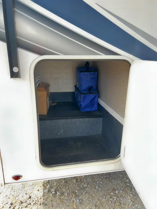 An open storage compartment of an RV rental, revealing two blue cooler bags, a cardboard box, and a textured floor.. Nexus Triumph 23T 2020