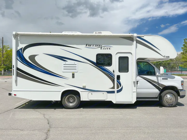 Bright white Class C RV rental with blue and black graphics, parked on asphalt under a cloudy sky. Ready for travel!. Thor Motor Coach Freedom Elite 2018