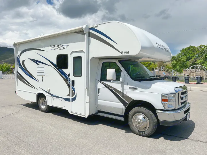 White Freedom Elite Class C RV rental, a motorhome with blue/black decals, parked on asphalt under cloudy skies.. Thor Motor Coach Freedom Elite 2018