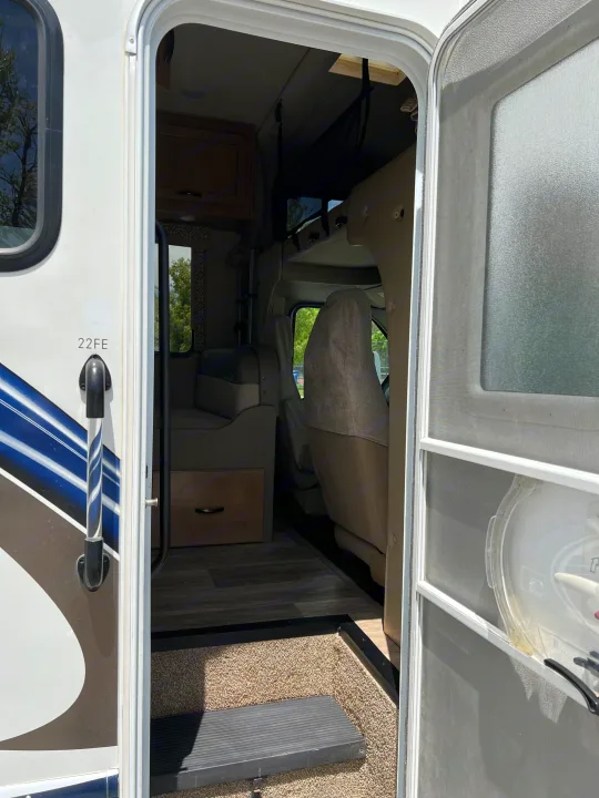 Open RV entrance showing exterior stripes, a screen door, and interior with driver's cabin, bunk bed, and living area. For an RV rental.. Thor Motor Coach Freedom Elite 2018