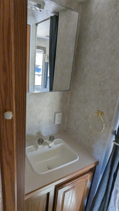 RV rental bathroom vanity featuring a white sink, mirror, wall outlet, and golden towel ring, set against light patterned walls and wood cabinetry.. Forest River Wildwood 2005