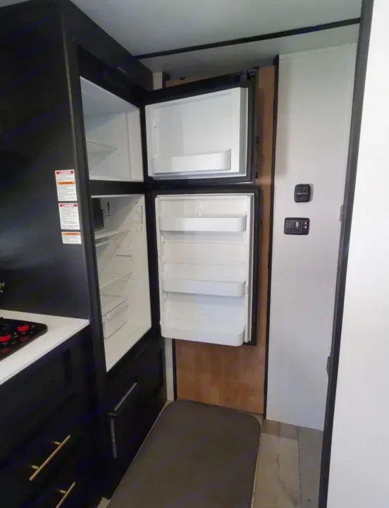 Inside an RV rental, a compact two-door refrigerator is open, revealing empty shelves. A stovetop and control panels are also visible.. Keystone RV Springdale 2022