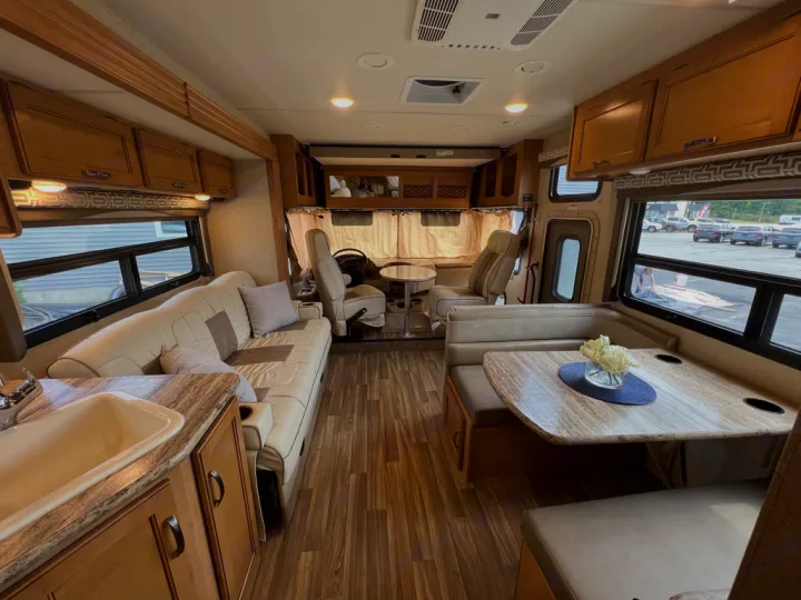 Inside a modern RV rental: open living space with light-colored sofa, dinette, & kitchen counter. Wood floors, ample light.. Thor Motor Coach A.C.E 2017