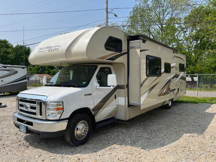 View of a beige and white Class C rv rental, featuring an extended slide-out, parked on a sunny gravel lot.. Thor Motor Coach Freedom Elite 2017
