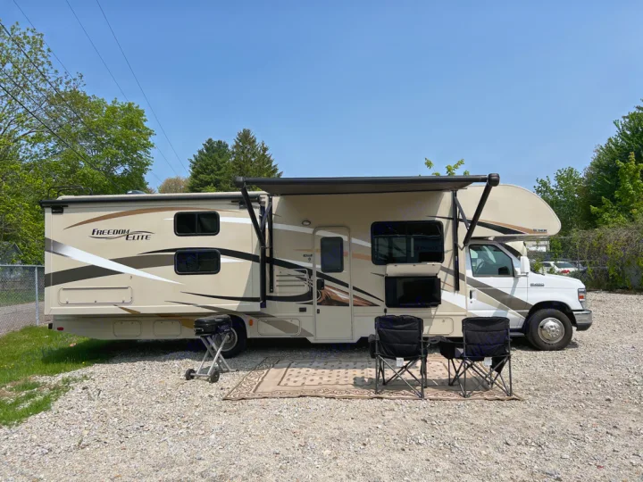 This **RV rental** is perfectly set up with an extended awning, outdoor TV, grill, chairs, and rug for camping on a gravel lot.. Thor Motor Coach Freedom Elite 2017