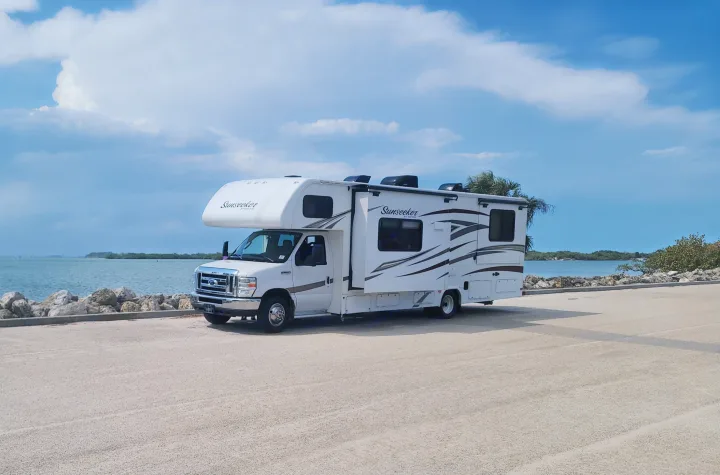 White Sunseeker RV rental parked on a paved lot by a calm bay, with rocky shoreline and distant islands.. Forest River Sunseeker 2017