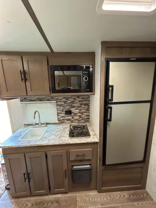 Modern RV rental kitchen: compact fridge, microwave, sink, two-burner stove, sleek gray cabinets, and light countertop.. Gulf Stream Ameri-Lite Super Lite 2021