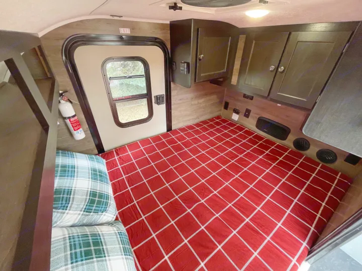 Cozy RV rental interior: a bed with red plaid bedding, overhead cabinets, an exit door with a window, and a fire extinguisher.. Braxton Creek Bushwhacker 2022