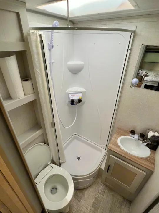 Compact RV rental bathroom features a corner shower, toilet, sink vanity, and built-in shelving, optimizing space.. No Boundaries 16 2023