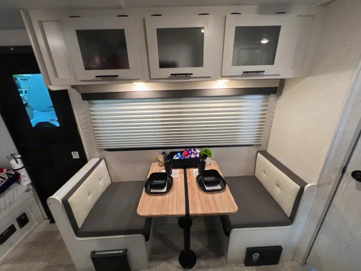 Modern RV rental dinette featuring a light wood table, grey cushioned benches, and overhead storage, ready for dining.. No Boundaries 16 2023