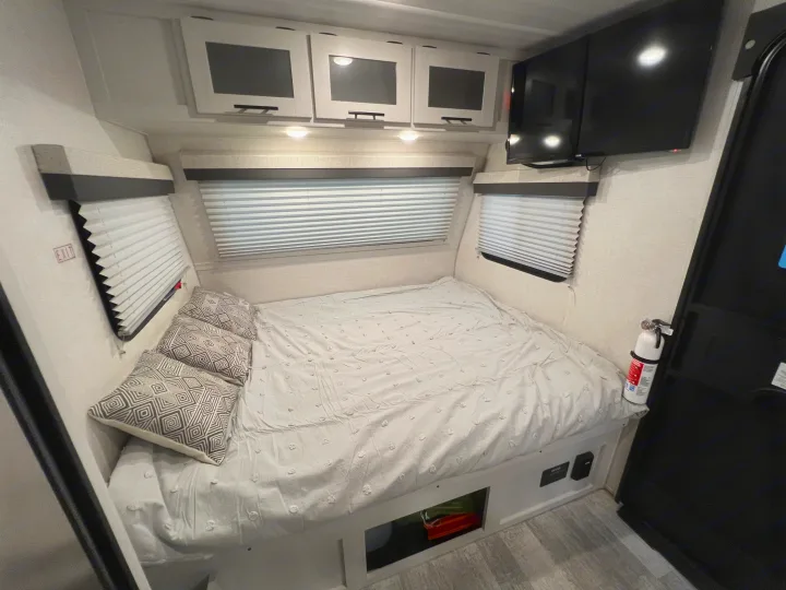 Cozy RV rental bedroom: a bed with storage, overhead cabinets, mounted TV, and fire extinguisher, ready for your travels.. No Boundaries 16 2023