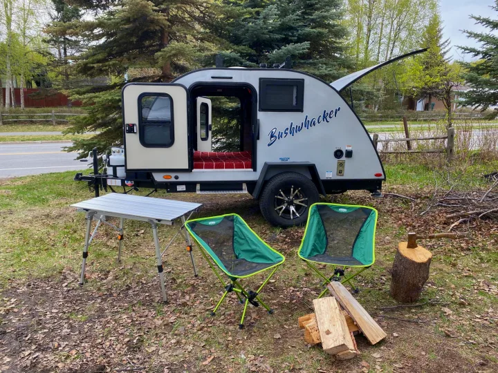 Bushwhacker teardrop RV rental with open doors, featuring a cozy interior. Outdoor table, chairs, and firewood ready for camping adventure.. Braxton Creek Bushwhacker 2022