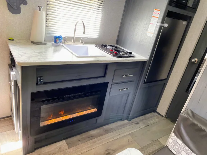 Modern RV rental kitchen featuring a sink, two-burner stove, mini-fridge, and a cozy electric fireplace with faux flames.. Dutchmen Aspen Trail 2021