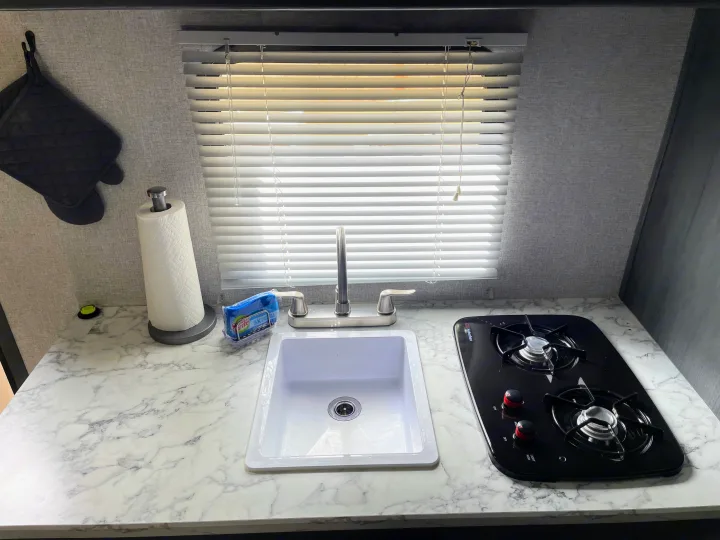 This RV rental kitchen features a countertop with a white sink, chrome faucet, two-burner stovetop, paper towels, and hanging pot holders. Window with blinds.. Dutchmen Aspen Trail 2021
