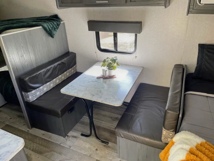 Inside an RV rental: Compact living space with a dinette, marble-look table, dark benches, and wood flooring. A cozy bunk is visible.. Dutchmen Aspen Trail 2021